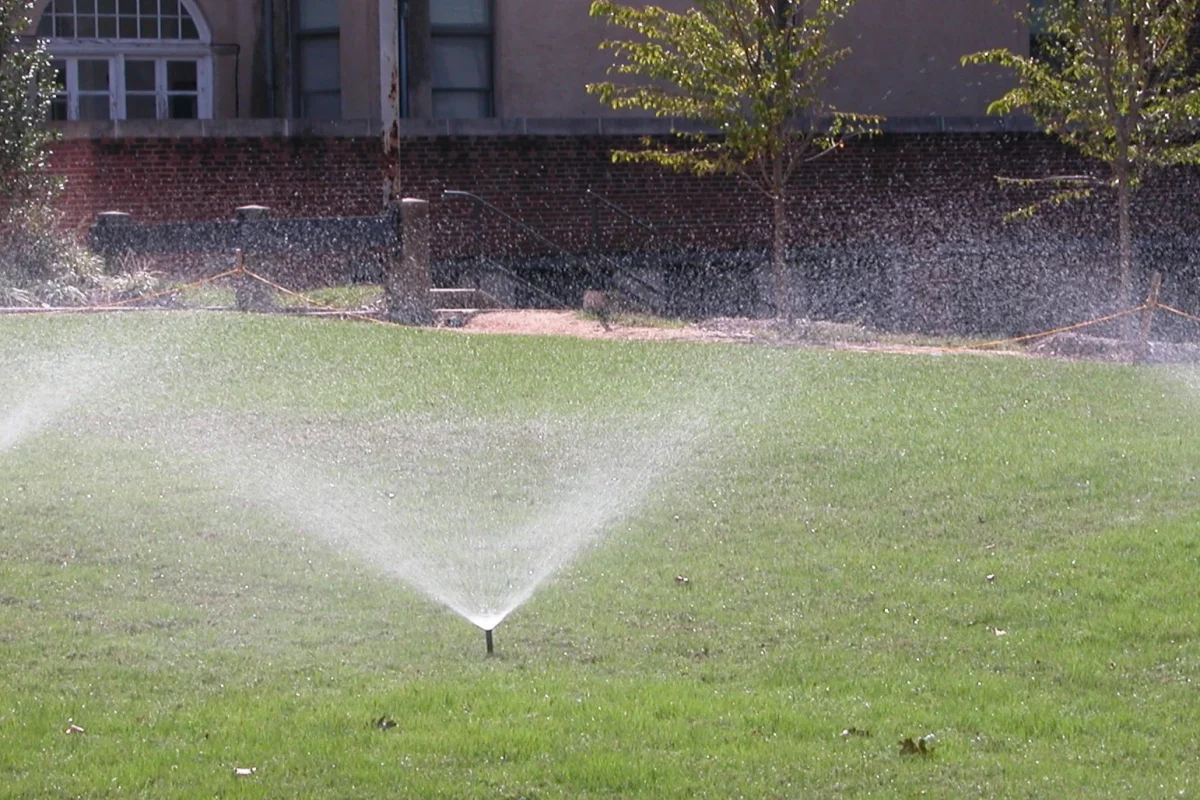 Three sprinklers spray water over a green lawn in front of a building, with trees, benches, and a brick wall in the background.